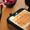 A close-up of a waffle maker with a waffle inside, with a bowl of raspberries in the background. Voir les images du produit