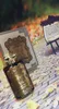 A perfume bottle and its box are displayed outdoors on a surface resembling a brick path, with fallen autumn leaves scattered around. A small easel with a document is also visible in the background. Produktbild-Vorschau