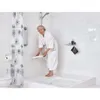 An elderly woman in a white bathrobe is in a bathroom, holding a white folding shower seat over a bathtub. Náhled obrázku produktu