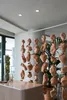 A collection of hanging ceramic vases in various abstract shapes and sizes, suspended from the ceiling in a modern interior space. Some are clustered around a potted plant, while others are arranged in vertical lines. A counter with a light-colored top and Produktbild-Vorschau