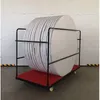A round folding table is stored vertically on a cart with a red base. The background is a white garage door. Produktbild-Vorschau