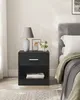 A black bedside table with a drawer and open shelf, next to a bed with a grey headboard and white bedding. Náhled obrázku produktu