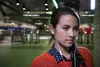 A young woman with earbuds in her ears, wearing an orange sweater and a blue polka-dot shirt, stands in a subway station. Anteprima dell’immagine prodotto