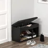 A black shoe cabinet with an open lid, showing shoes inside, placed next to a white door. Produktbild-Vorschau