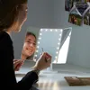 A young woman is applying makeup in front of a lighted makeup mirror. Produktbild-Vorschau