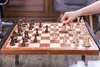 A person's hand is shown moving a white chess piece on a wooden chessboard. Podgląd zdjęcia produktu