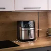 An air fryer is placed on a kitchen countertop, with cabinets above and a stovetop to the left. Dimensions are indicated with lines and text. Produktbild-Vorschau