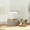A round storage ottoman with a lid sits on a circular rug, flanked by a pair of tan cowboy boots. A tall white vase with branches is in the background. Produktbild-Vorschau