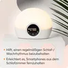 A white and beige Lumie light alarm clock is displayed on a white surface, with a plant in the background. Text describing the product's benefits is present. Produktbild-Vorschau