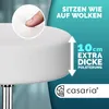 Close-up of a white stool with text overlays highlighting comfort and padding. A finger is shown pressing into the cushion, demonstrating its softness. The brand logo is also visible. Produktbild-Vorschau