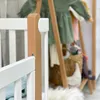 Close-up of a white baby crib with a natural wood accent, with blurred baby clothes and a basket in the background. Produktbild-Vorschau