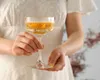 A person in a white dress holds a champagne coupe filled with a golden liquid. Náhľad obrázku produktu