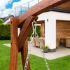 A close-up view of a wooden swing set frame with metal chains hanging from it, set in a garden with a house in the background. Náhled obrázku produktu