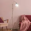 A floor lamp is shown in a living room setting next to a chair and a sofa. Produktbild-Vorschau