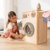 A young girl is playing with a wooden toy washing machine, which is part of a larger play set that includes a clothesline, ironing board, and laundry basket. The scene is set in a bright, domestic environment. Produktbild-Vorschau