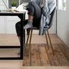 A person is sitting at a desk, writing in a notebook, with a laptop and a plant nearby. The floor has a wood-plank design. Produktbild-Vorschau