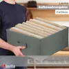 A person is holding a gray fabric storage box filled with folded clothes. The background shows a blurred interior setting. Produktbild-Vorschau