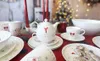 A Christmas-themed table setting with plates, cups, a teapot, candles, and cookies, with a blurred Christmas tree in the background. Produktbild-Vorschau