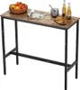 A narrow, long bar table with a brown wood-grain top and black metal legs, set against a white background. It is decorated with a basket of fruit, a magazine, a glass of ice, and two bottles of beer. Produktbild-Vorschau