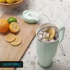 A mint green insulated tumbler filled with ice and a slice of orange, placed on a countertop next to a wooden cutting board with oranges, limes, and lemon slices. A light green lid and a silver straw are also visible. Anteprima dell’immagine prodotto