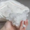 A hand holding a handful of white pillow stuffing, with a partially opened pillow in the background. Náhled obrázku produktu