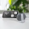 A smartphone displaying smart home controls and a wireless charger on a white surface with a blurred green plant in the background. Anteprima dell’immagine prodotto