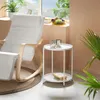 A white side table with two books and two decorative cups on top, placed next to a white rocking chair in a room with natural light coming from a window. Náhled obrázku produktu