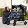 A woman is interacting with two babies in a double stroller outdoors. Produktbild-Vorschau