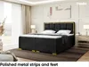 A bedroom scene featuring a black upholstered boxspring bed with a gold-trimmed headboard and gold feet, set against a neutral background with curtains and a window. Produktbild-Vorschau