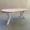 A modern oval dining table with unique white legs against a textured wall and concrete floor. Voir les images du produit