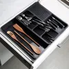 A black cutlery organizer drawer insert filled with silverware and wooden kitchen utensils is shown within an open kitchen drawer. Náhled obrázku produktu