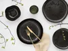 A flat lay of black stoneware plates and a bowl, with gold cutlery and a small green glass, arranged with delicate branches with green leaves on a white background. Náhľad obrázku produktu