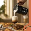 A person is pouring frothed milk from a black milk frother into a glass of coffee, creating a latte or cappuccino. There are also pastries and coffee beans on a wooden board in the foreground. Náhled obrázku produktu