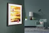A framed picture of California with a cyclist on a beach at sunset hangs on a wall above a teal cabinet with a lamp, a decorative horse, and a teal armchair with a blanket and pillow. Produktbild-Vorschau