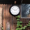A round outdoor clock with a thermometer and hygrometer is mounted on a rustic wooden fence, with green vines growing around it. Náhled obrázku produktu