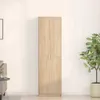 A tall, narrow wardrobe with two doors stands against a plain white wall. A potted plant is visible to the right, and a small side table with a sofa is partially visible to the left. Produktbild-Vorschau