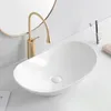 A white oval vessel sink with a gold faucet on a white countertop, with a round mirror in the background. Náhľad obrázku produktu
