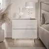 A white, glossy nightstand with two drawers and chrome legs, placed next to a bed. It is decorated with a bust sculpture and a lamp. Produktbild-Vorschau