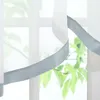 A close-up view of a sheer curtain with a gray satin ribbon trim, hanging in front of a window with green foliage visible outside. Produktbild-Vorschau