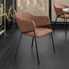 A brown upholstered chair with a black metal frame is shown in a room with a dark wooden floor and a large window overlooking a cityscape. Produktbild-Vorschau