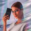 A woman with dramatic makeup holds a black smartphone in front of a shimmering, iridescent background. Produktbild-Vorschau