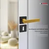 A close-up of a black and gold door handle set on a door, with a blurred background of a room interior. Produktbild-Vorschau