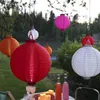 Several paper lanterns of various colors are hanging from tree branches in an outdoor setting, with a table set with drinks and candles in the foreground. Podgląd zdjęcia produktu