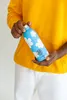 A person wearing a yellow hoodie and white pants holds a blue water bottle with white cloud patterns. Produktbild-Vorschau