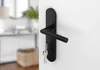 A black door handle with keys attached, mounted on a white door. The background shows a blurred interior scene. Produktbild-Vorschau
