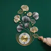 A decorative wall clock with a ginkgo leaf design is mounted on a green wall. A hand is shown adjusting one of the leaves. Produktbild-Vorschau