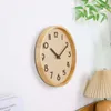 A wooden wall clock with dark brown numbers and hands is mounted on a white wall. A lamp with a pleated shade and a plant with green leaves are visible in the background. Produktbild-Vorschau