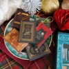 A collection of miniature book ornaments, resembling classic literature, arranged on a decorative plate with festive Christmas decorations in the background. Náhled obrázku produktu