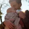 A father is feeding his baby from a bottle while holding the baby in a carrier. Produktbild-Vorschau