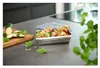 A stainless steel food container filled with chopped vegetables and herbs, placed on a kitchen counter next to a cutting board with sliced zucchini and basil. Produktbild-Vorschau
